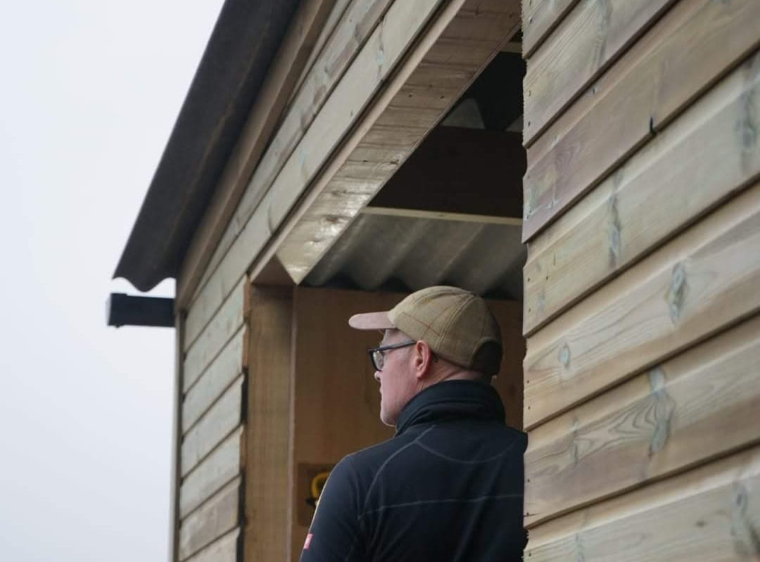 A close-up of a builder in a baseball cap looking at a timber-clad farm building, next to text about getting a quote for farm buildings.