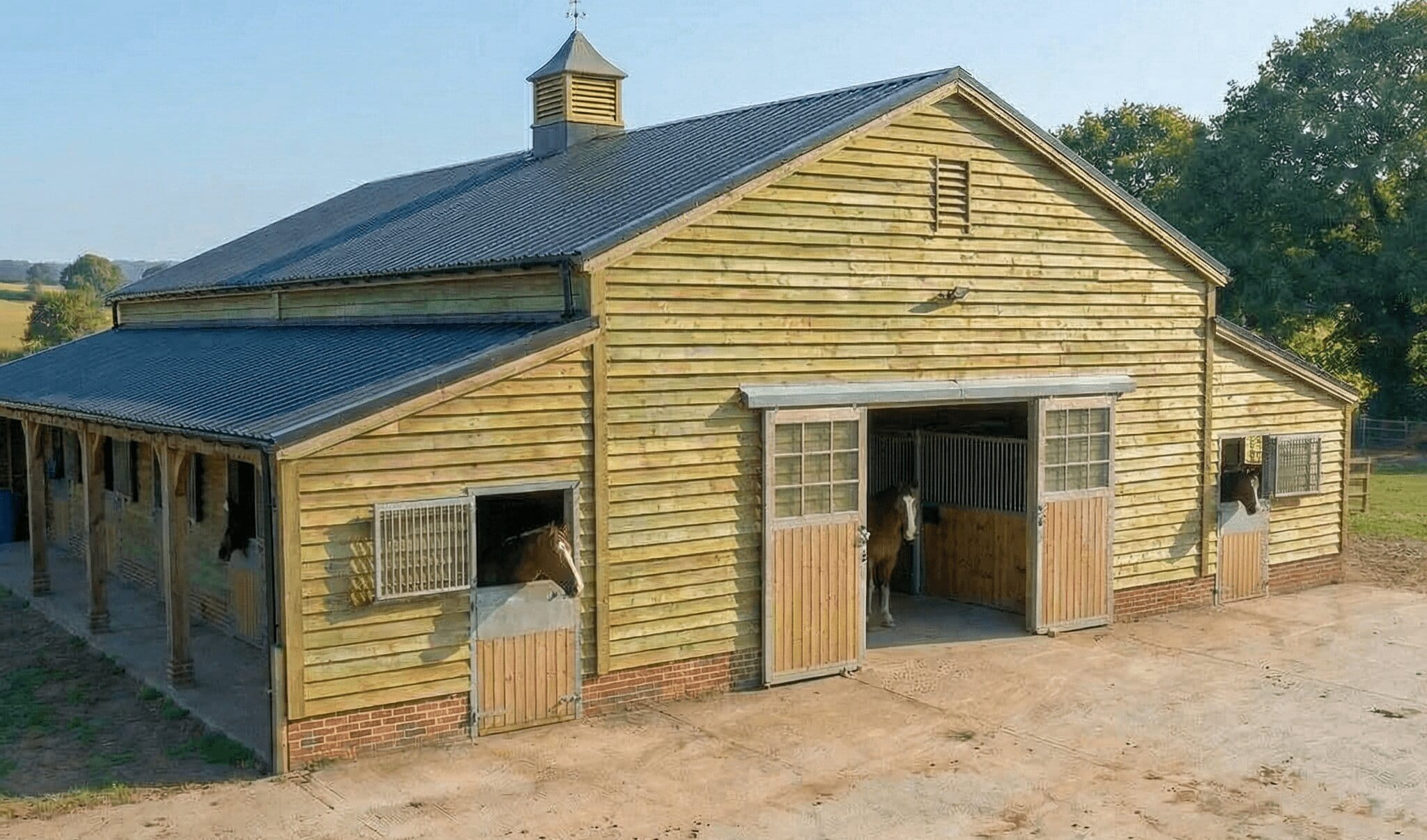 A large timber American Barn with a central aisle, sliding doors, and a decorative roof cupola, housing horses in an all-in-one sheltered facility.