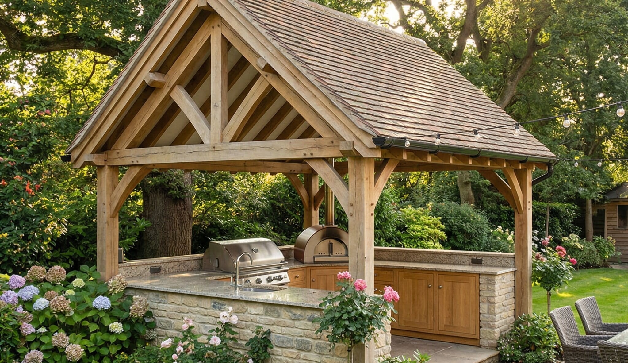 A luxury oak-framed outdoor kitchen shelter with a tiled roof covering a built-in BBQ, pizza oven, and granite countertops in a garden.