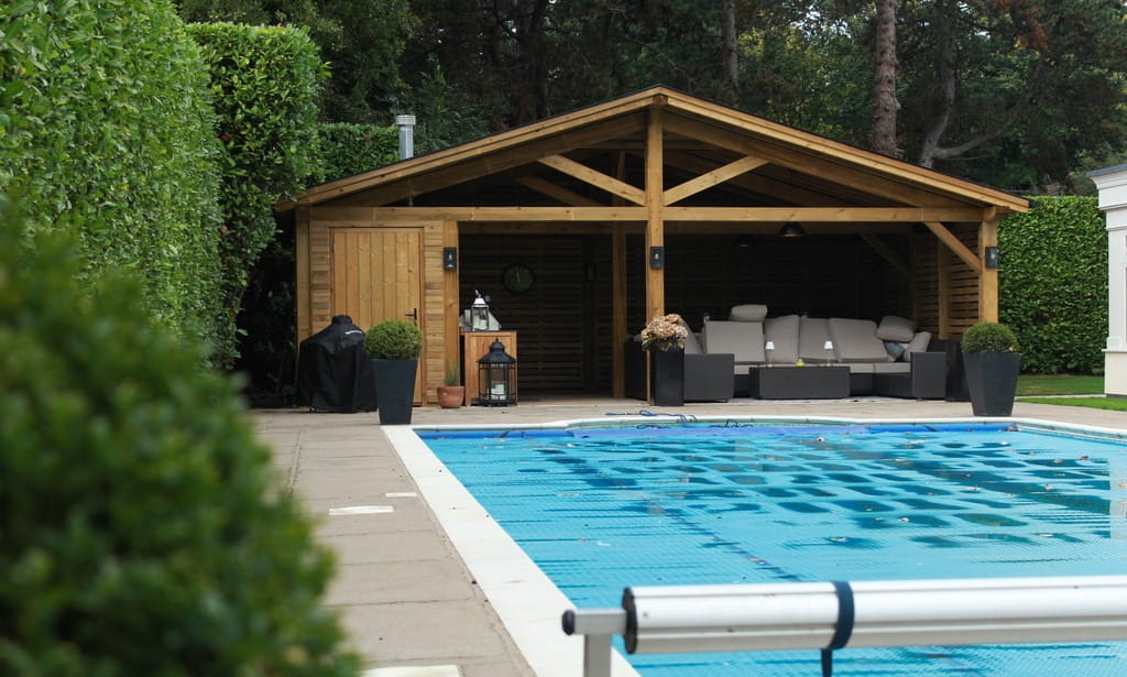 A luxury hand-crafted timber outdoor living structure with a seating area, located next to a blue swimming pool and surrounded by privacy hedging.