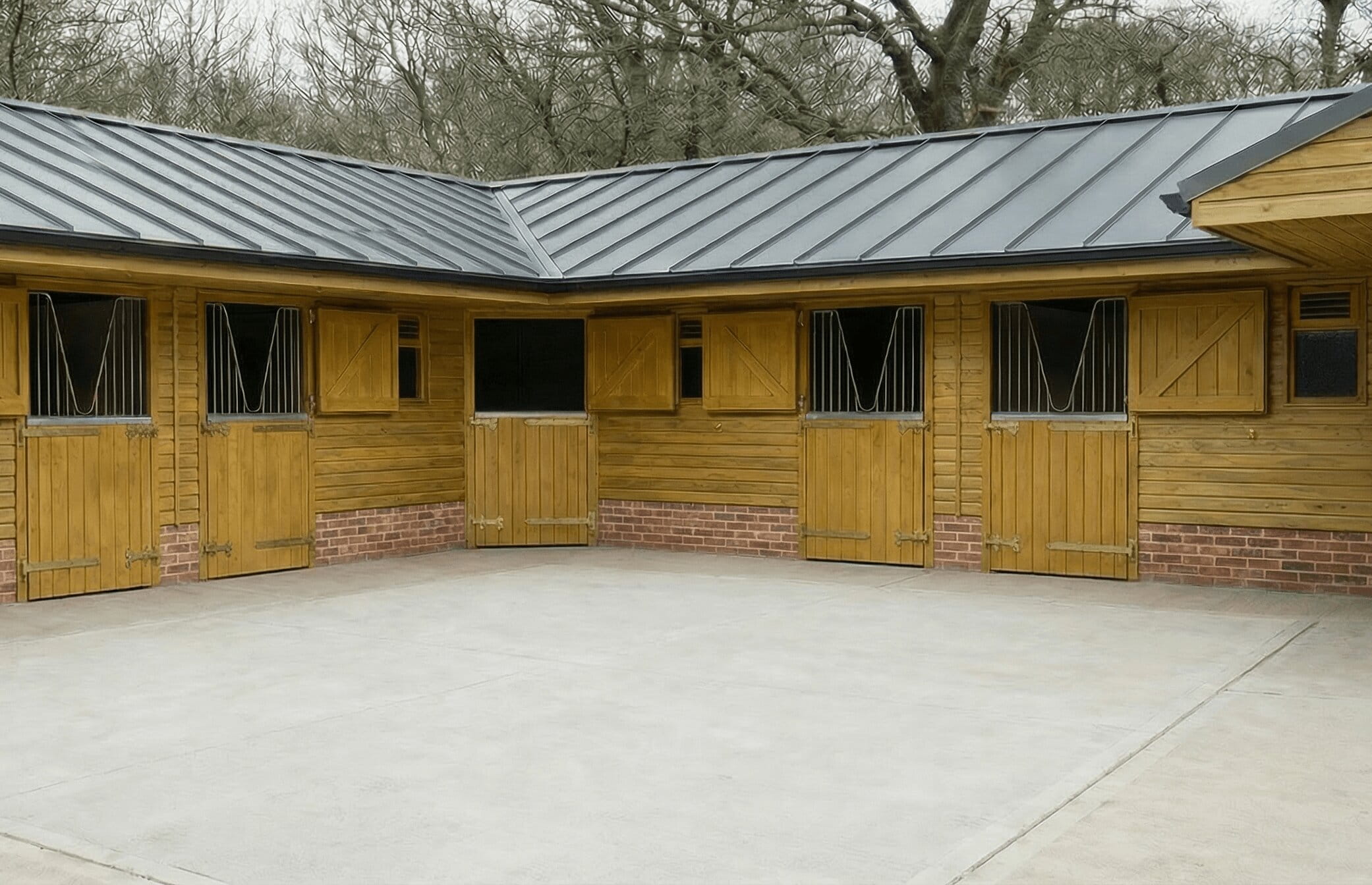 An L-shaped timber stable block with tanalised cladding, a dark grey metal profile roof, and brick plinth walls surrounding a concrete courtyard.