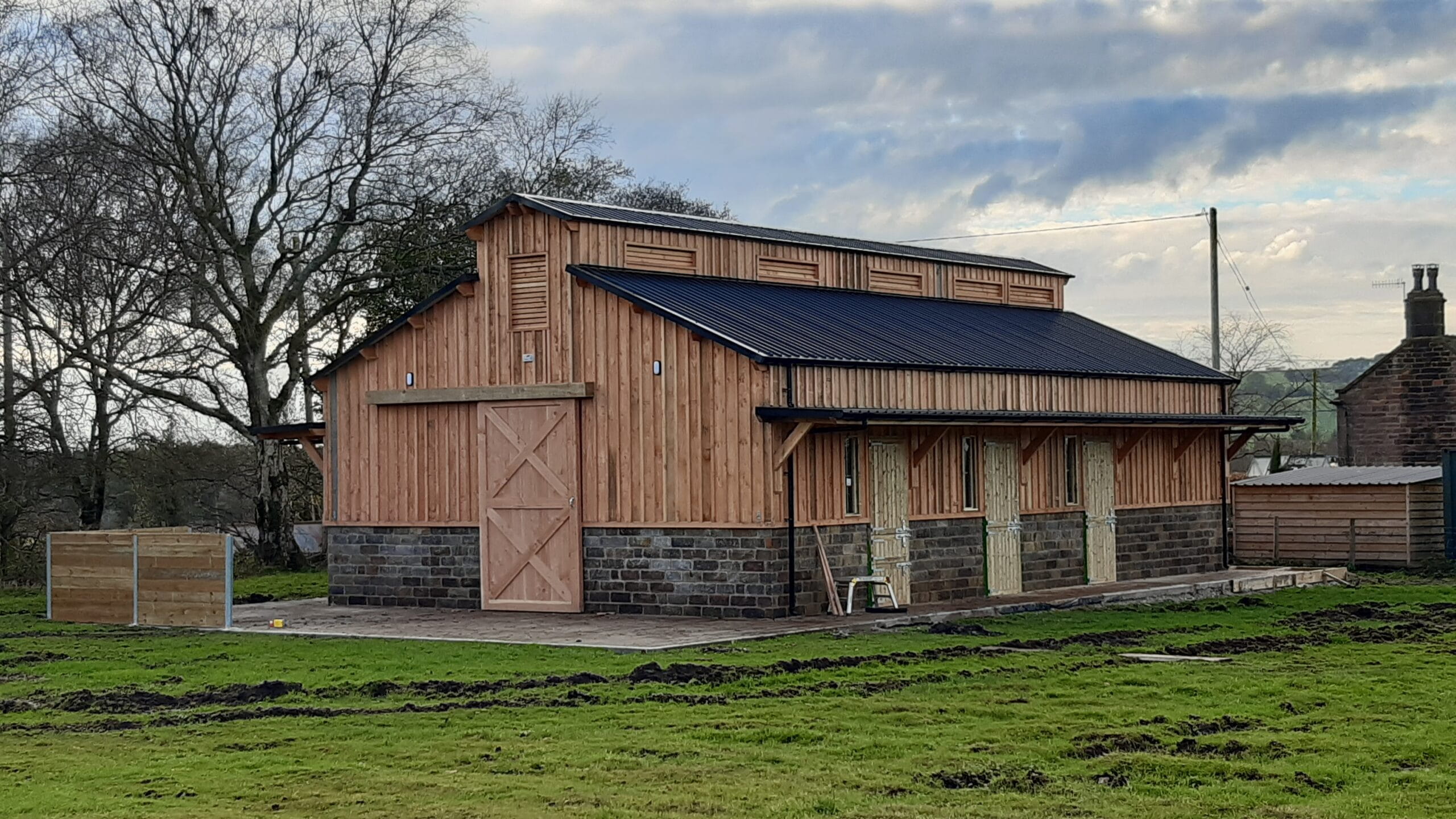 A side view of a large timber-framed barn with multiple stable doors, a stone base, and a black corrugated roof under a cloudy sky.