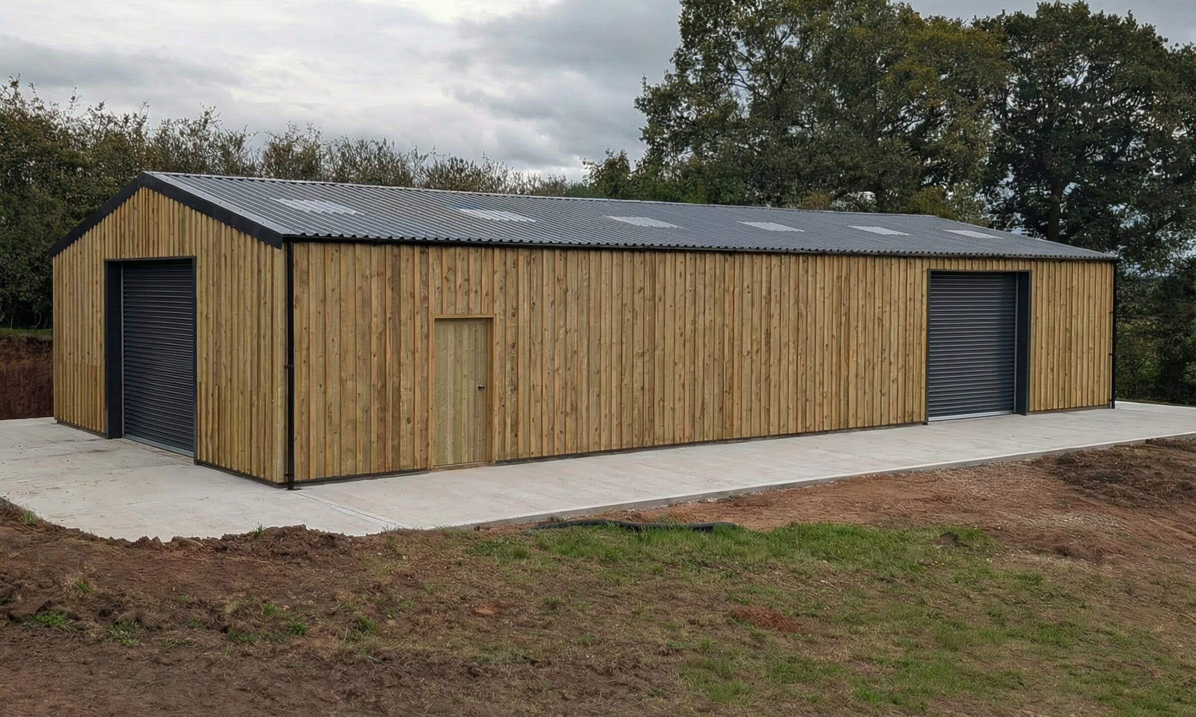 A large, modern timber-clad agricultural building with a grey corrugated metal roof and black roller shutter doors on a concrete base.