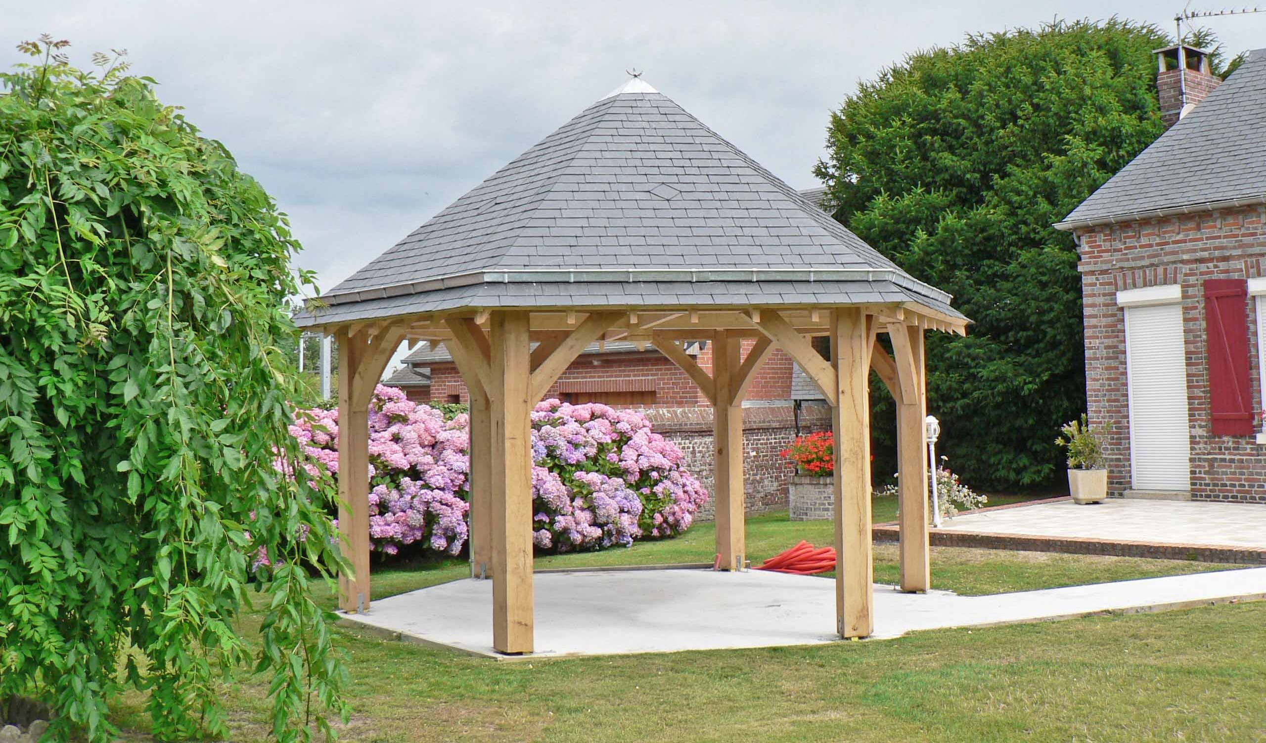 A hand-crafted oak framed timber gazebo with a grey tiled roof standing in a garden next to pink hydrangea bushes.