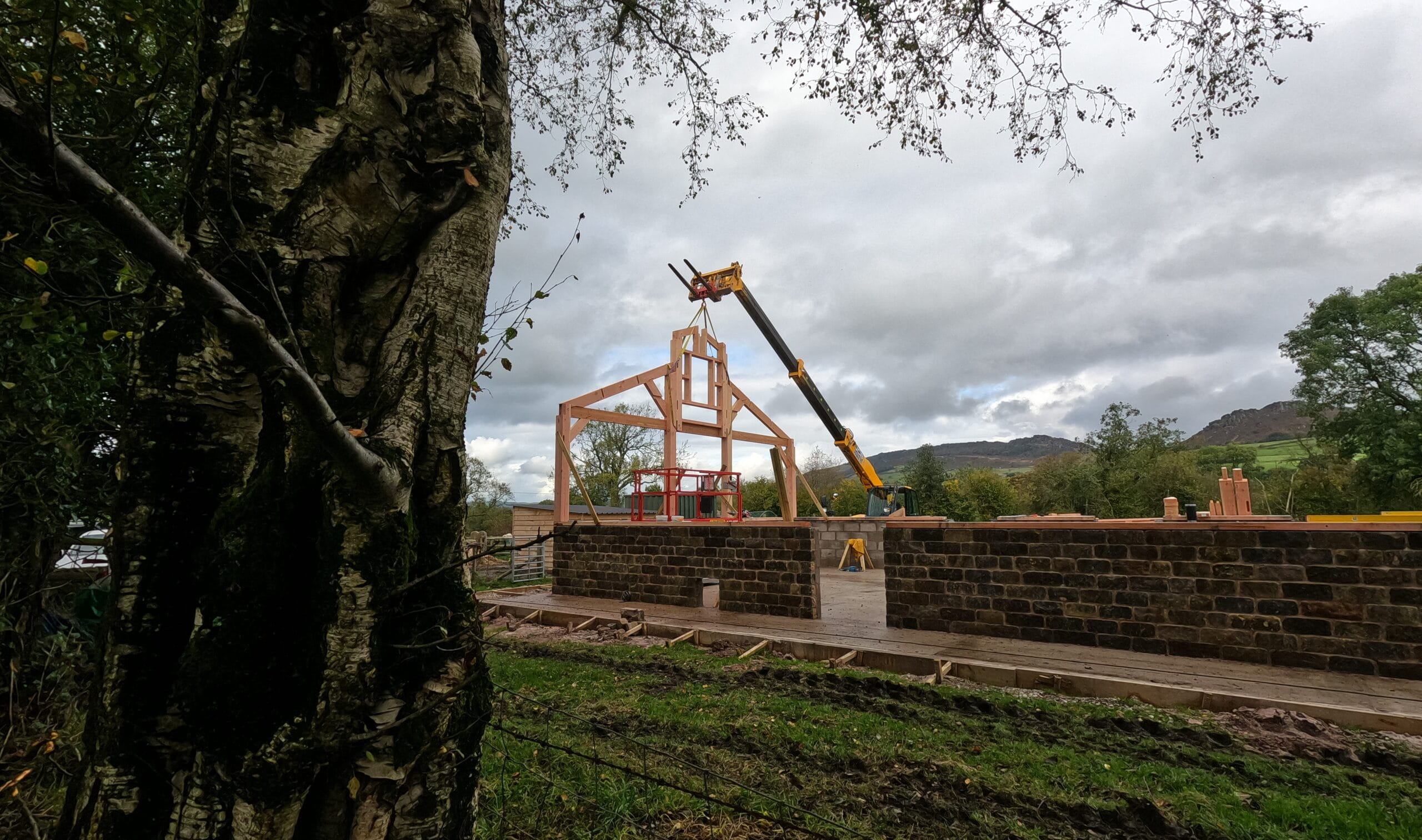A telehandler crane lifting a large timber gable frame onto a stone-walled foundation at a rural construction site under a cloudy sky.