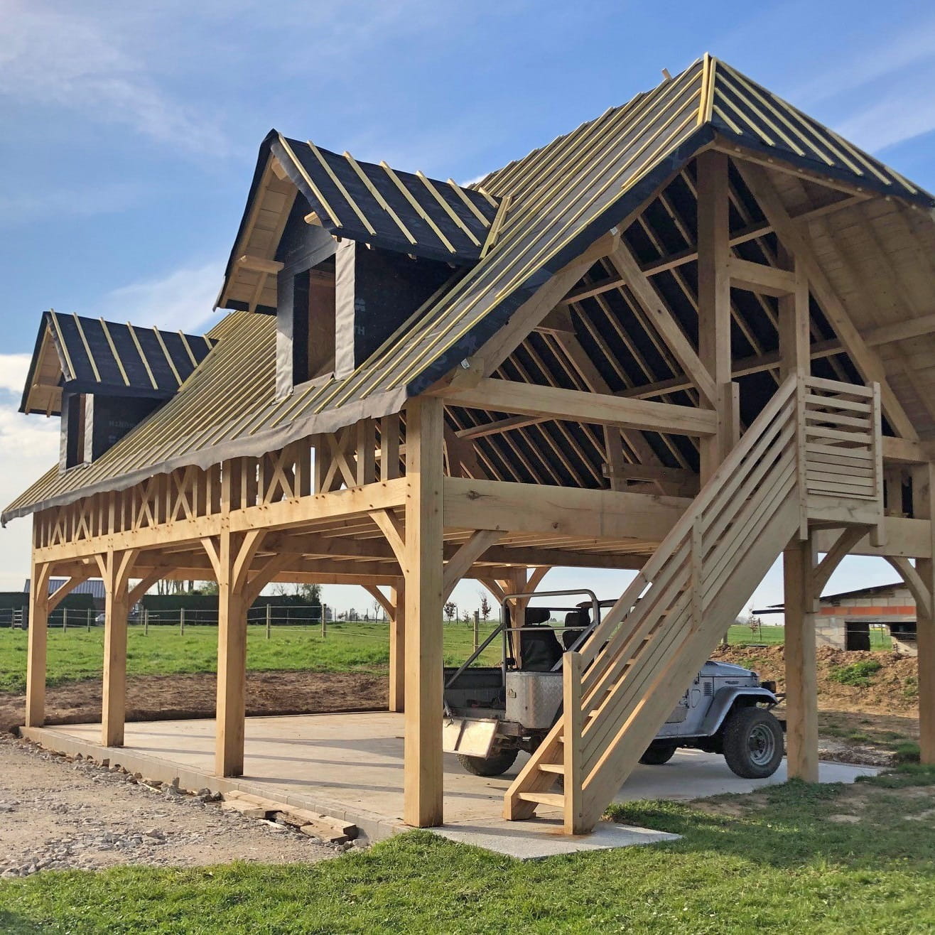An oak-framed two-story garage or garden room under construction, showing the skeletal timber frame and external staircase.