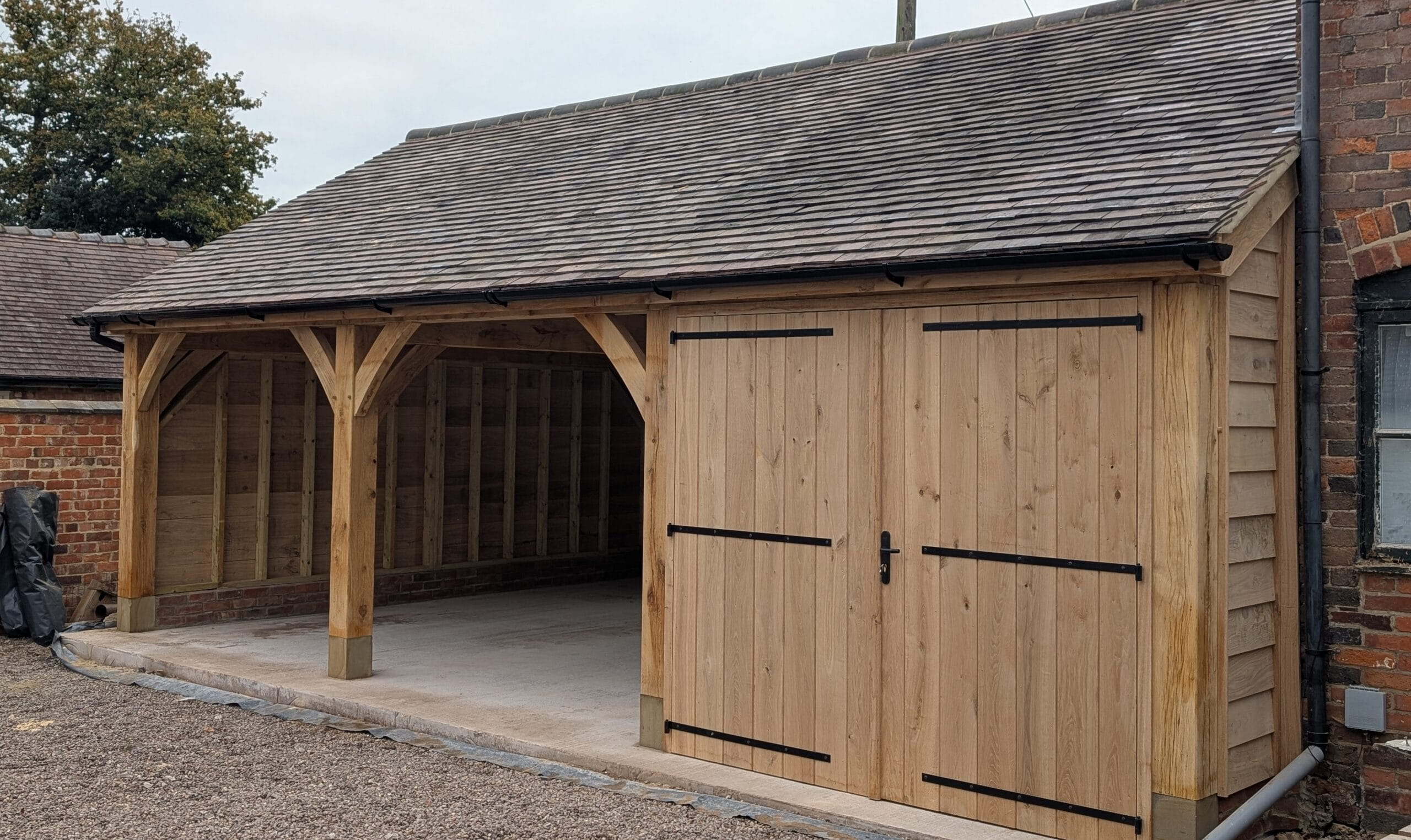A completed oak-framed triple-bay building featuring two open carports and one enclosed workshop with large timber double doors and black ironmongery.