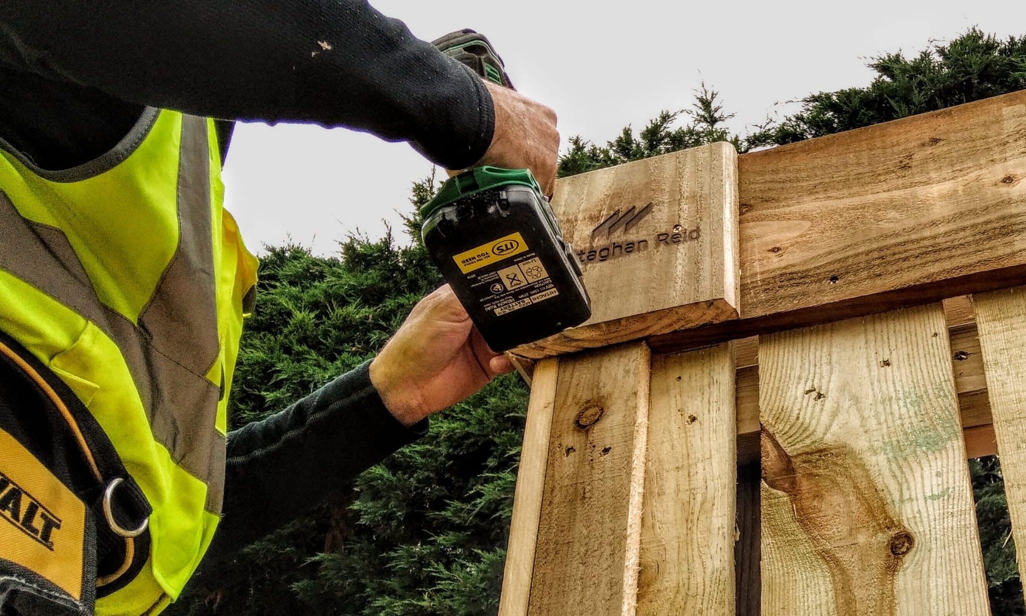 A close-up of a carpenter in a high-visibility vest using a power drill to secure a timber component featuring an engraved Mottaghan Reid logo.
