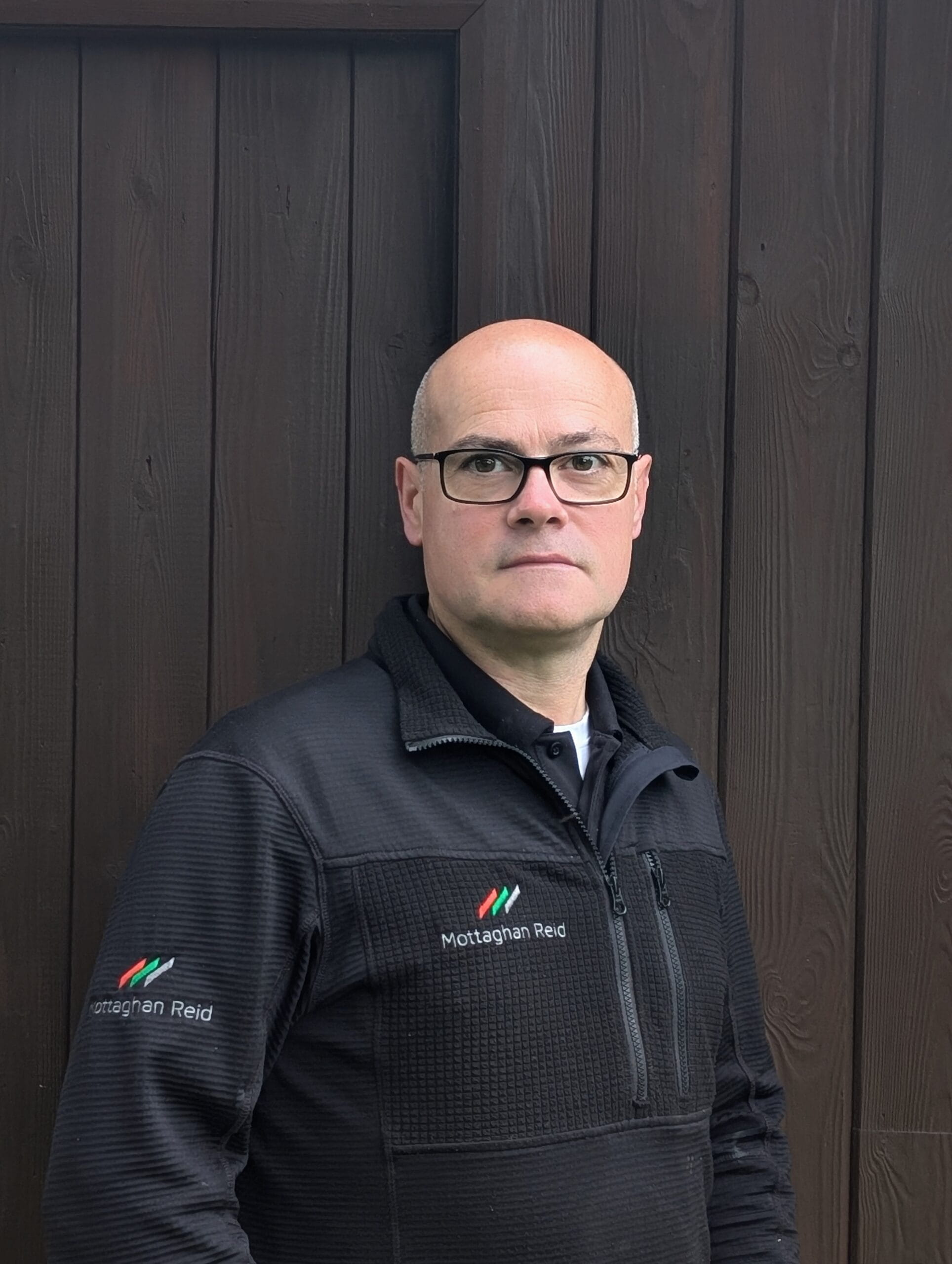 A portrait of Russel Godden, Owner and Site Carpenter of Mottaghan Reid, wearing a company-branded black fleece jacket against a dark wood-paneled wall.