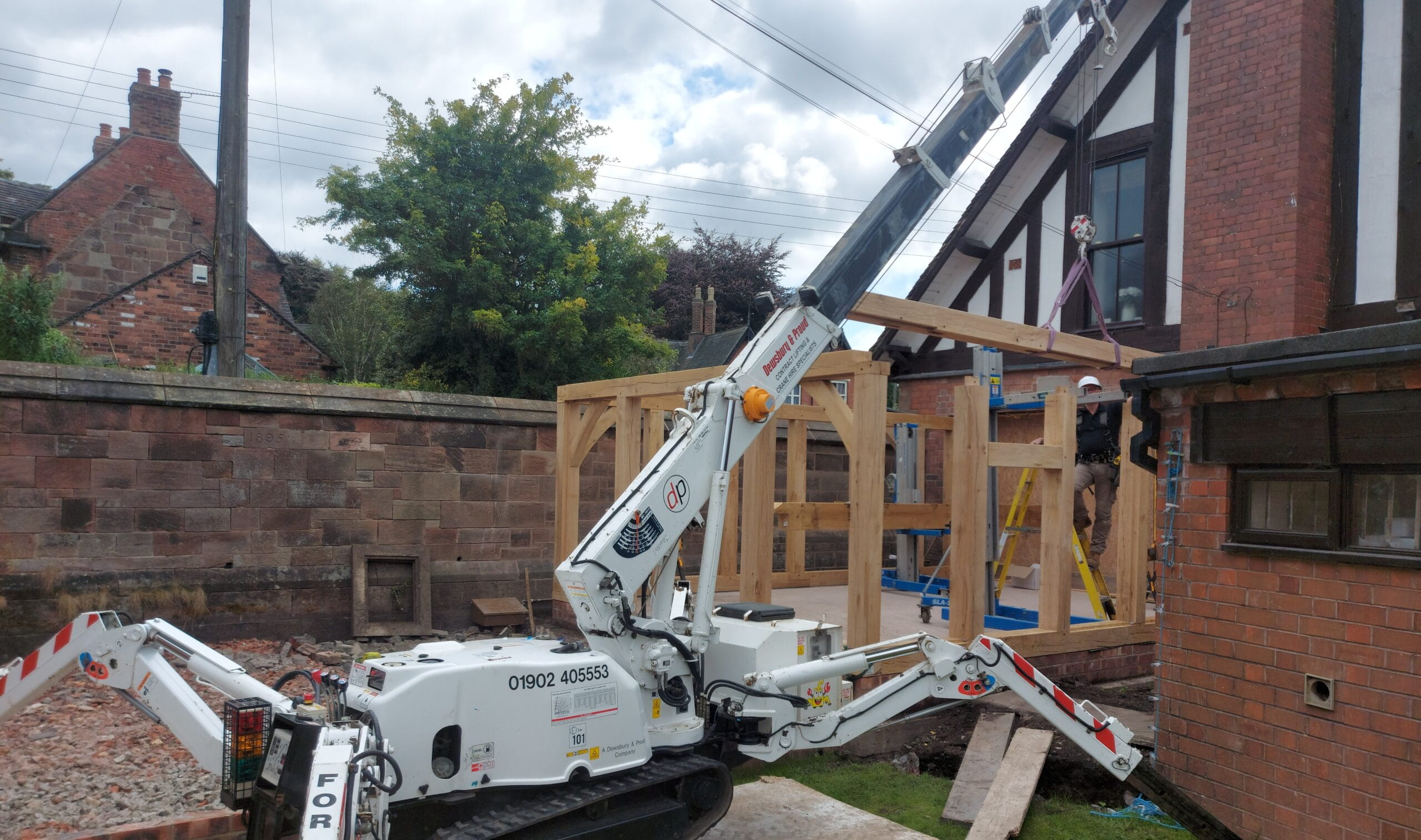 A white spider crane lifting a heavy timber beam into place for a precision-cut oak frame assembly on a residential construction site.